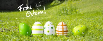Fünf dekorierte Ostereier in Grün und Weiß stehen auf Gras in einer sonnigen Umgebung im Freien und symbolisieren Frische und Naturstrom. Frohe Ostern! mit Hasenohren steht in weißer Schrift über den Eiern.